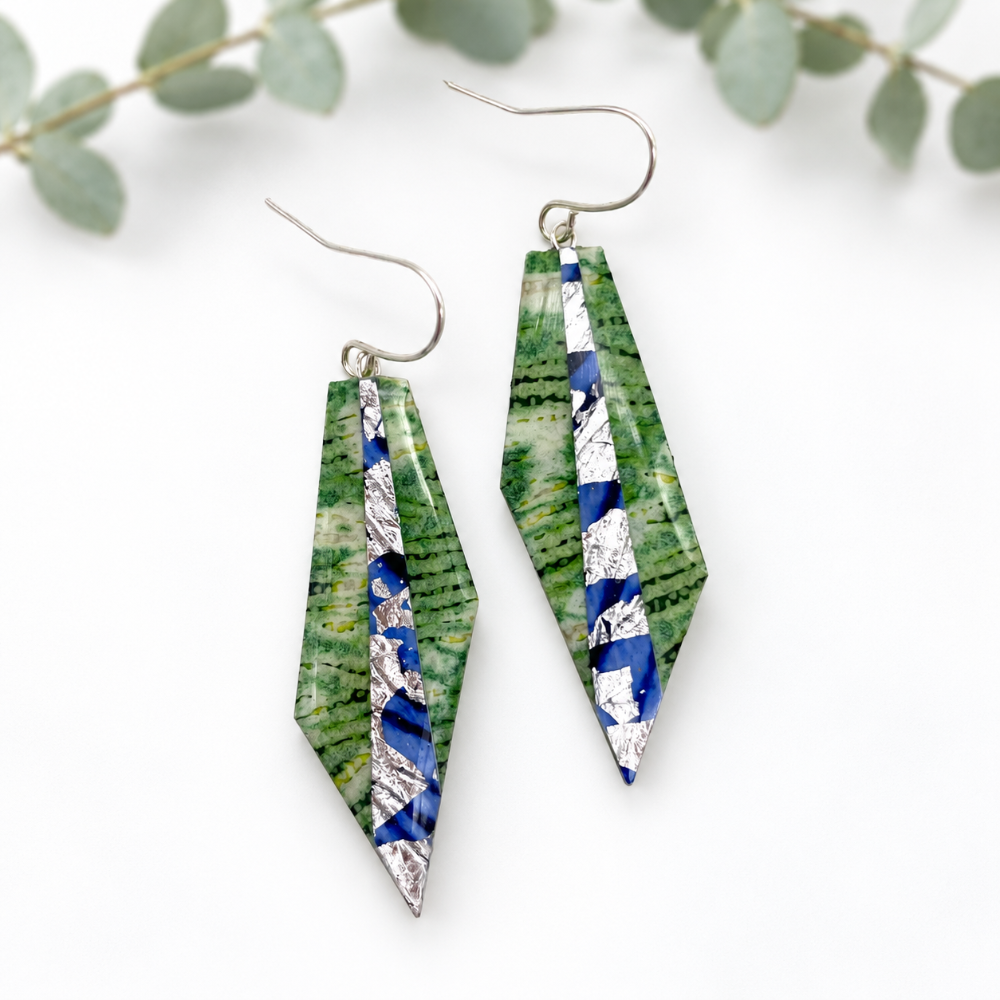 A pair of geometric drop earrings with green fern-patterned panels, blue and white striped sections, and silver fishhook wires, displayed on a clean white background with soft eucalyptus leaves in the background.