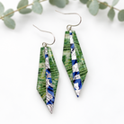 A pair of geometric drop earrings with green fern-patterned panels, blue and white striped sections, and silver fishhook wires, displayed on a clean white background with soft eucalyptus leaves in the background.