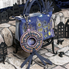 A blue handbag with a detailed illustration of gothic houses under a crescent moon, displayed on a small black stand. The bag features a large circular brooch charm with a central clock and hanging straps, set against a background of miniature black house cutouts and stone tiles.