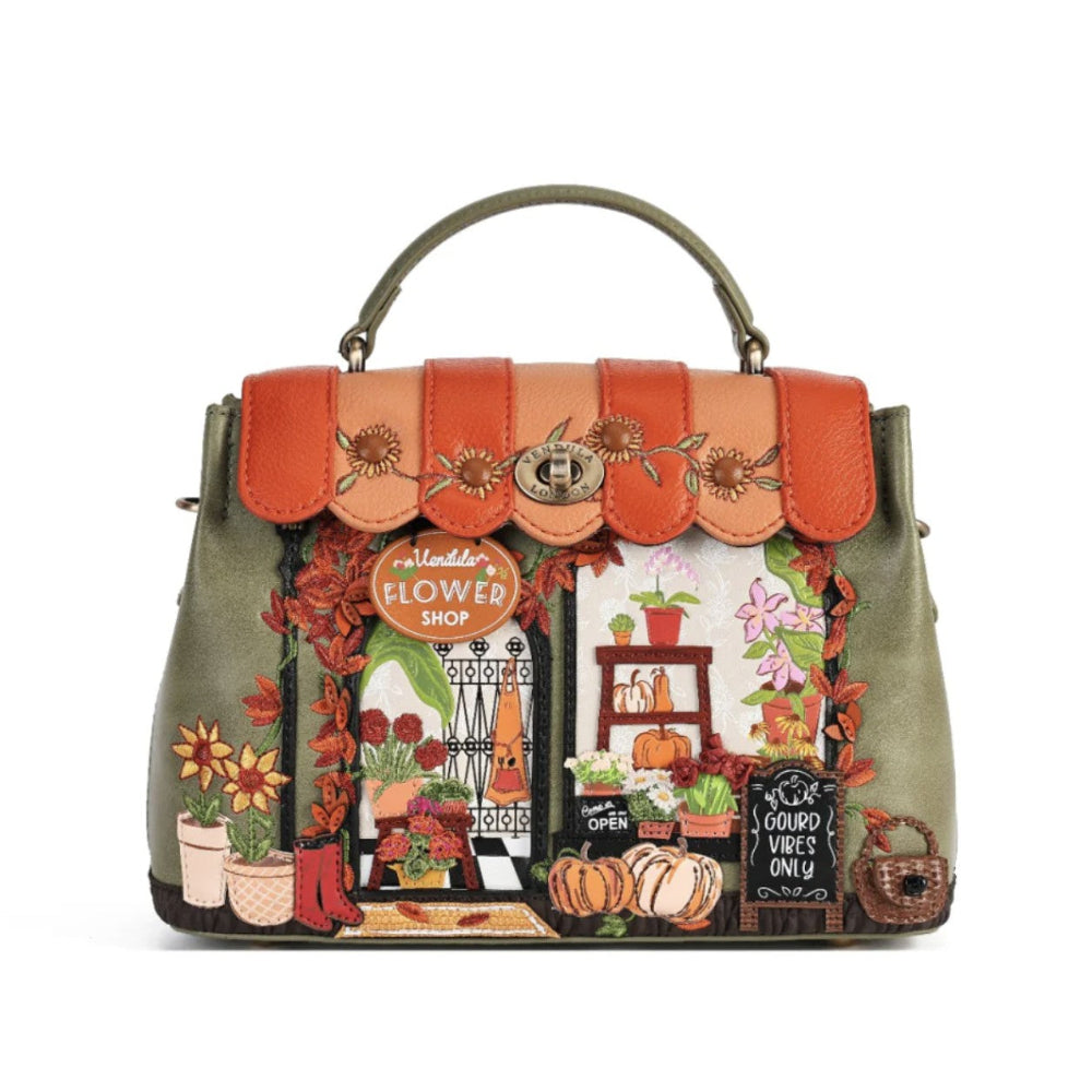 A structured handbag with a top handle, featuring a detailed autumn-themed illustration on the front depicting a flower shop entrance with pumpkins, flowers, and a sign that reads 'Vendula Flower Shop'. The bag has a sage green base, a peach and orange scalloped flap, and embroidered floral details.
