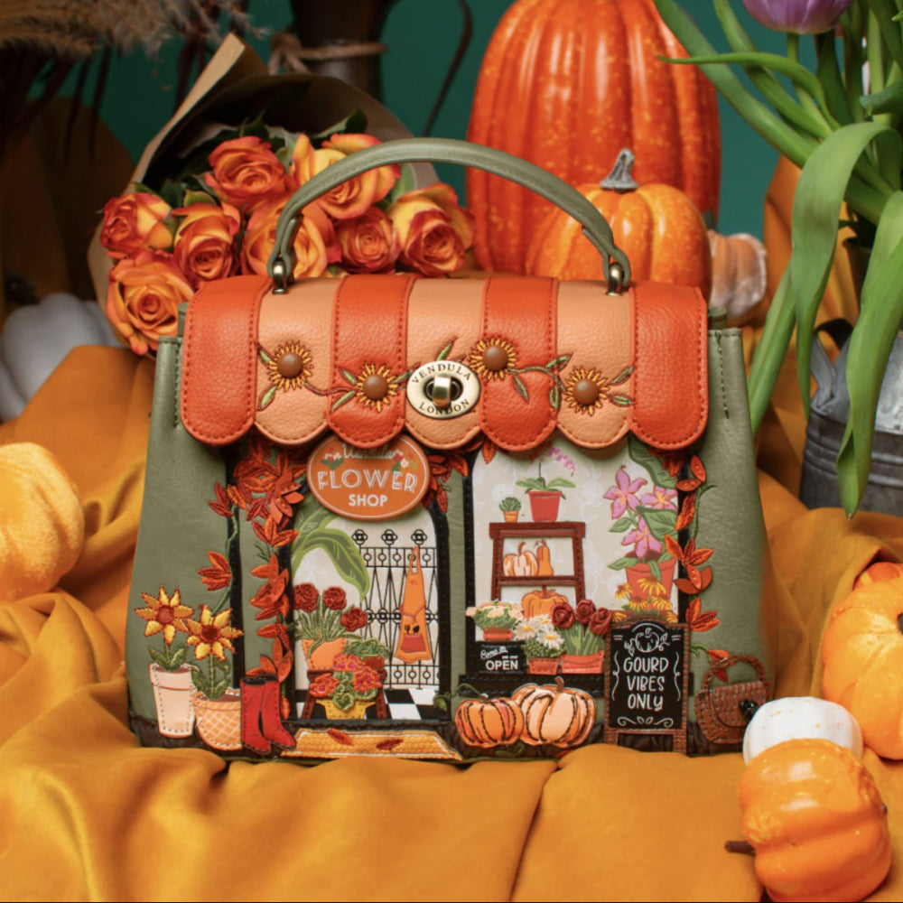 A decorative handbag styled in a fall-themed setting with pumpkins, roses, and draped orange fabric. The bag has a scalloped orange leather top flap with floral details, a green handle, and a front panel featuring autumn illustrations of flowers, pumpkins, and a storefront sign reading 'Flower Shop'.