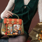 A woman holds a decorative handbag in a styled indoor setting with wicker baskets and autumn flowers. The bag has a sage green base with orange striped trim, floral and pumpkin illustrations, and a small top handle.