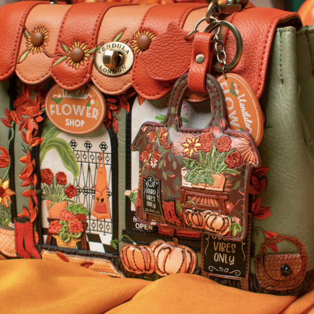 Close-up of a decorative handbag with a warm autumn floral design. The bag features layered orange and peach faux-leather flap details with sunflower embellishments, a gold-tone 'Vendula London' logo medallion, and a hanging charm shaped like a shopping bag filled with pumpkins, flowers, and fall-themed signs including 'Flower Shop' and 'Good Vibes Only'.