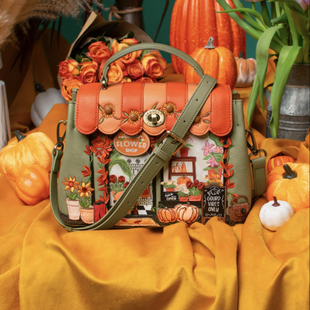 A decorative autumn-themed handbag displayed on draped mustard-yellow fabric with pumpkins, roses, and greenery in the background. The bag has an olive green body and strap, orange leather trim, floral and storefront embroidery, and a central gold-tone clasp.