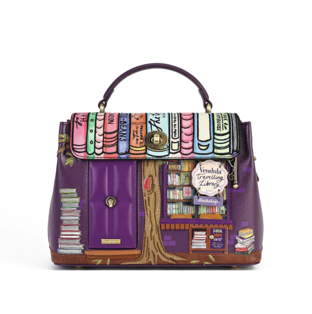 A purple handbag with a library-themed printed design on the front flap and body, featuring book spines, shelves, a tree trunk, and a sign reading 'Vendula Travelling Library'.