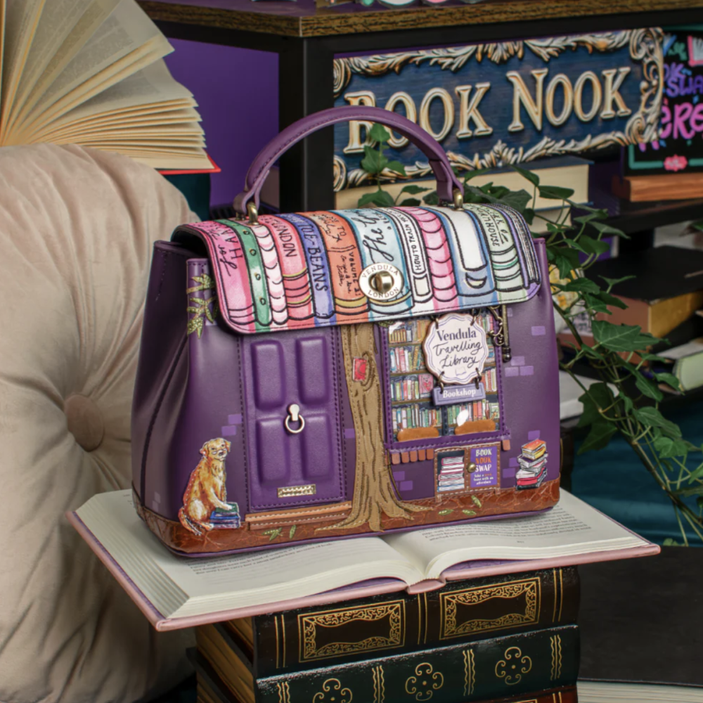 A purple handbag with a library-themed design, featuring illustrated book spines across the flap and a bookstore facade on the front, displayed on an open book in a cozy book-filled room.