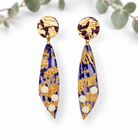 A pair of statement dangle earrings featuring a deep purple and gold marbled pattern with textured gold leaf details and white circular accents, displayed on a clean white background with soft eucalyptus leaves.