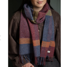 A person wearing a chunky woven scarf with large color-blocked stripes in dusty blue, burgundy, rust orange, and olive green, styled over a brown jacket.