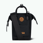 A black Cabaia shoulder bag with a top handle, adjustable shoulder strap, and two circular brown leather brand patches, shown against a plain white background.