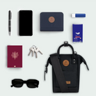 Collection of travel essentials including a black backpack, phone, passport, keys, sunglasses, and other items on a light gray background.