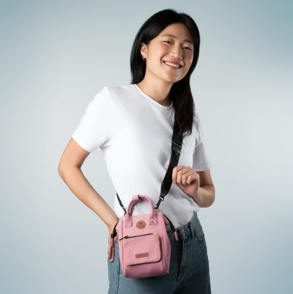 A woman wearing a white t-shirt and blue jeans models a small pink crossbody bag with a black strap against a light gray studio background.