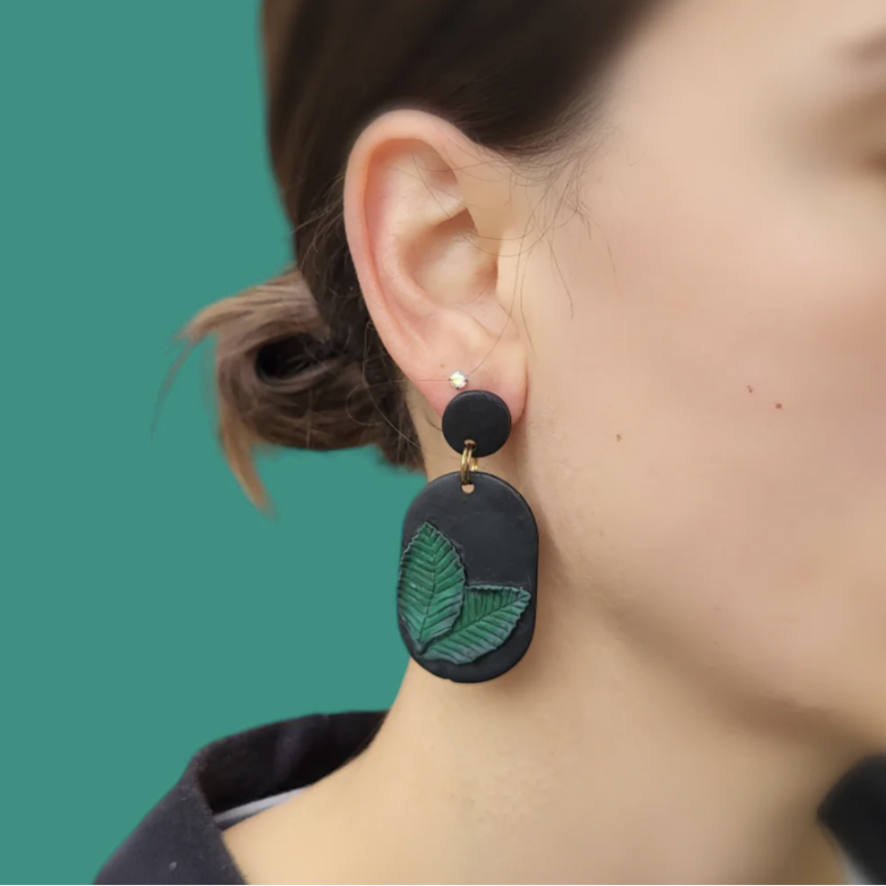 Black earrings with green leaf design worn by a person against a teal background