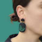 Black earrings with green leaf design worn by a person against a teal background
