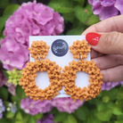 Orange floral earrings on a card held in front of purple flowers