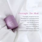 A lavender purple jar of Bare By Vogue Cosmetics Overnight Tan Mask resting on draped white fabric, with product details listed in pink and gray text on the right.