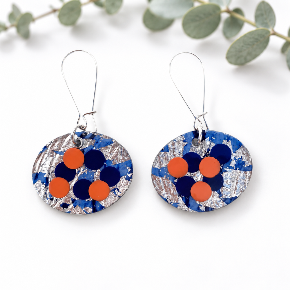 A pair of handmade oval drop earrings with a blue, silver, and orange abstract pattern, hanging from silver fishhook wires on a white background with eucalyptus leaves.