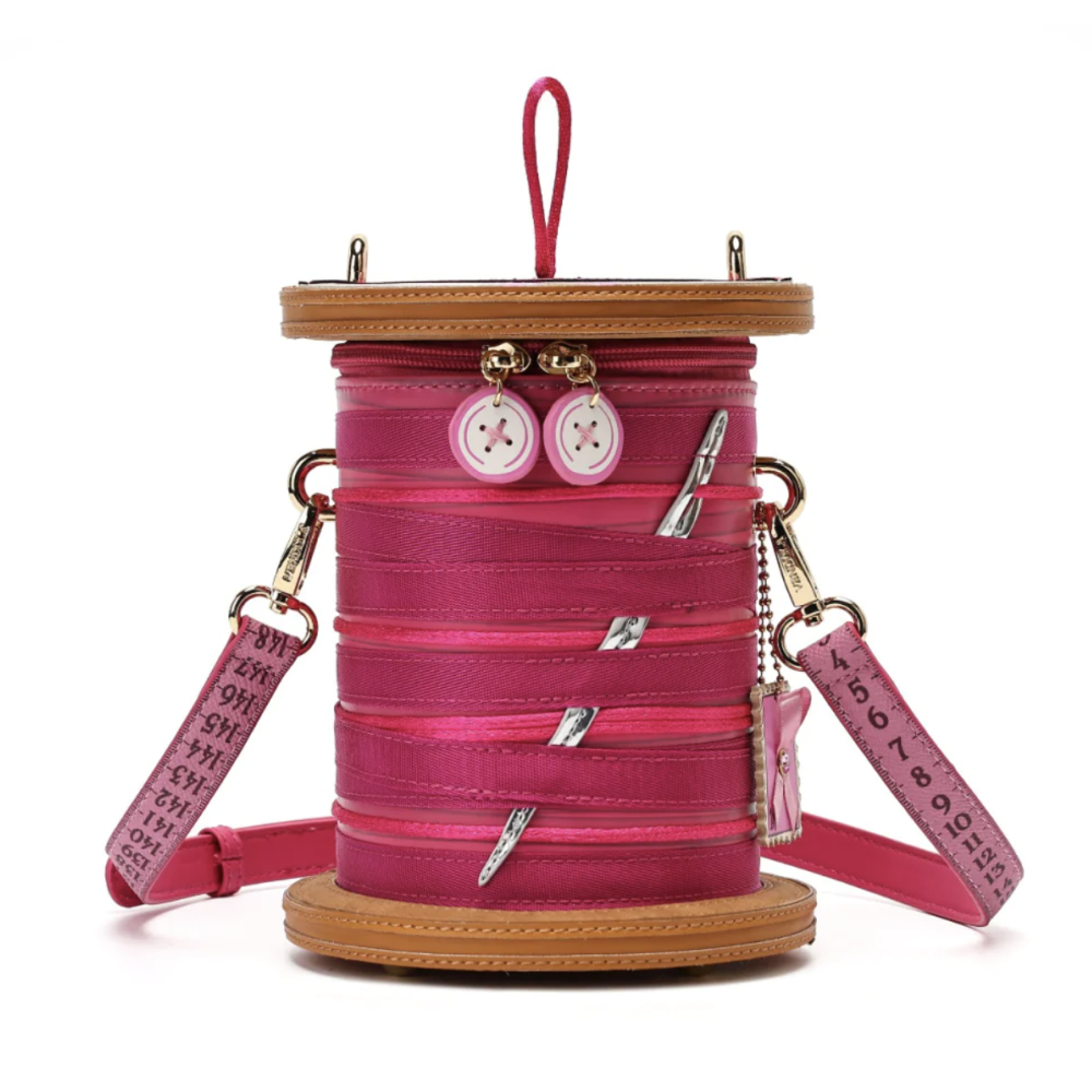 A pink spool-shaped shoulder bag with a tan faux-leather top and base, pink measuring tape straps, silver nail-shaped hardware, and two decorative buttons on the front, shown on a white background.