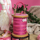 A pink spool-shaped handbag with a top handle, gold-tone hardware, and decorative buttons, styled on a small black stand against a pink background with mannequins, ribbon, and fabric props.