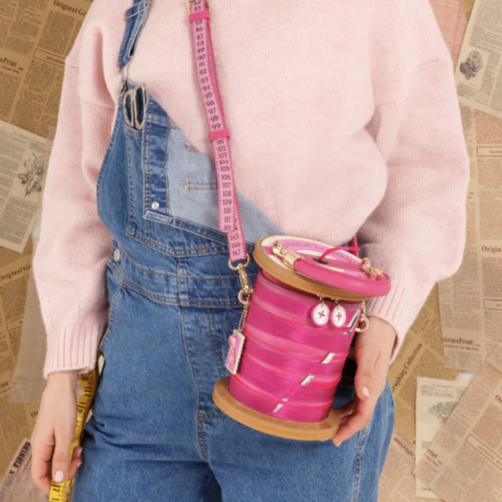 A person wearing a light pink sweater and blue denim overalls holds a bright pink spool-shaped handbag with a measuring tape strap against a background of newspaper pages.