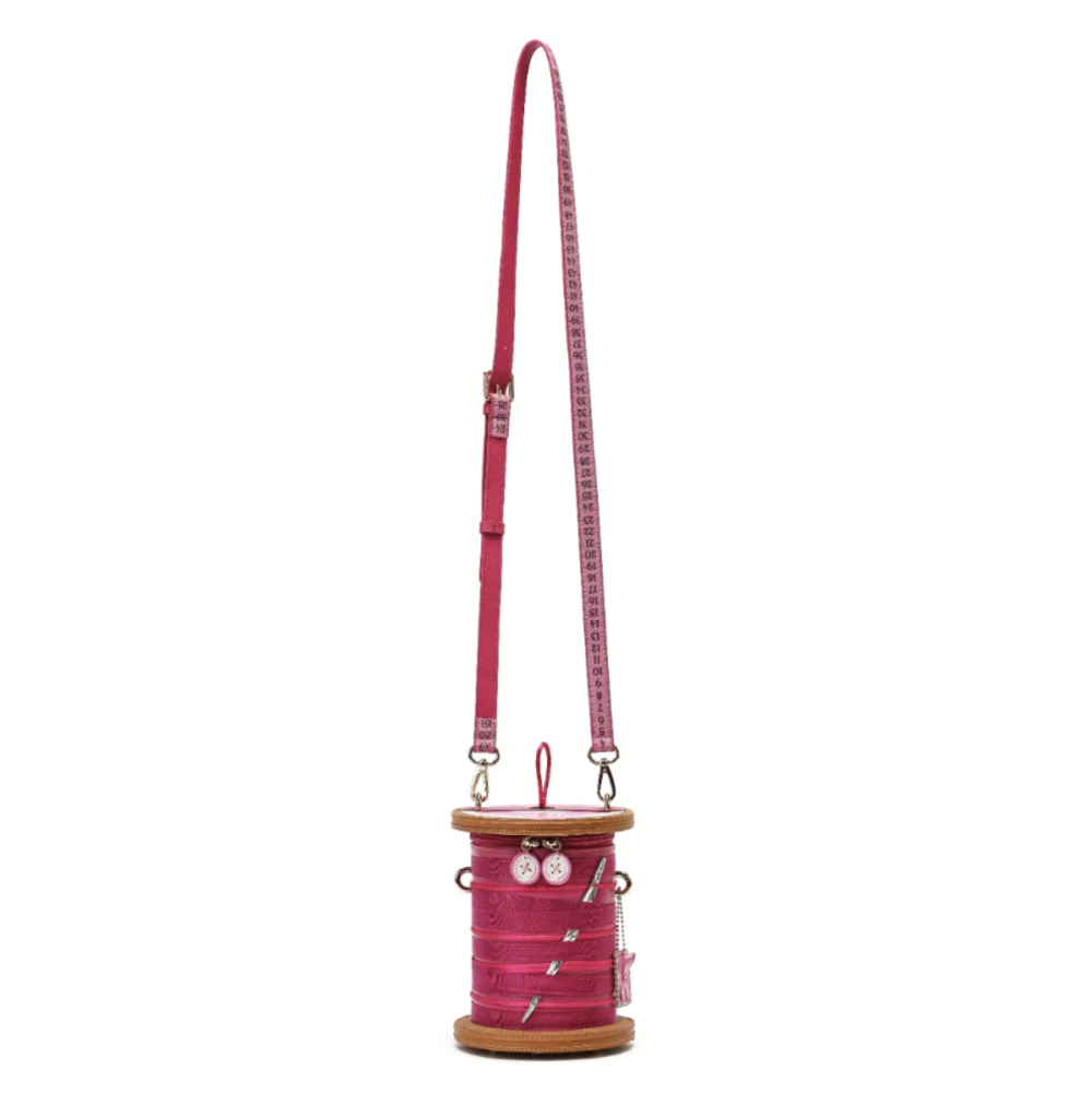 A pink spool-shaped crossbody bag with a long matching strap, featuring decorative buttons and pins on the spool, set against a plain white background.