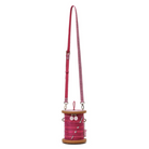 A pink spool-shaped crossbody bag with a long matching strap, featuring decorative buttons and pins on the spool, set against a plain white background.