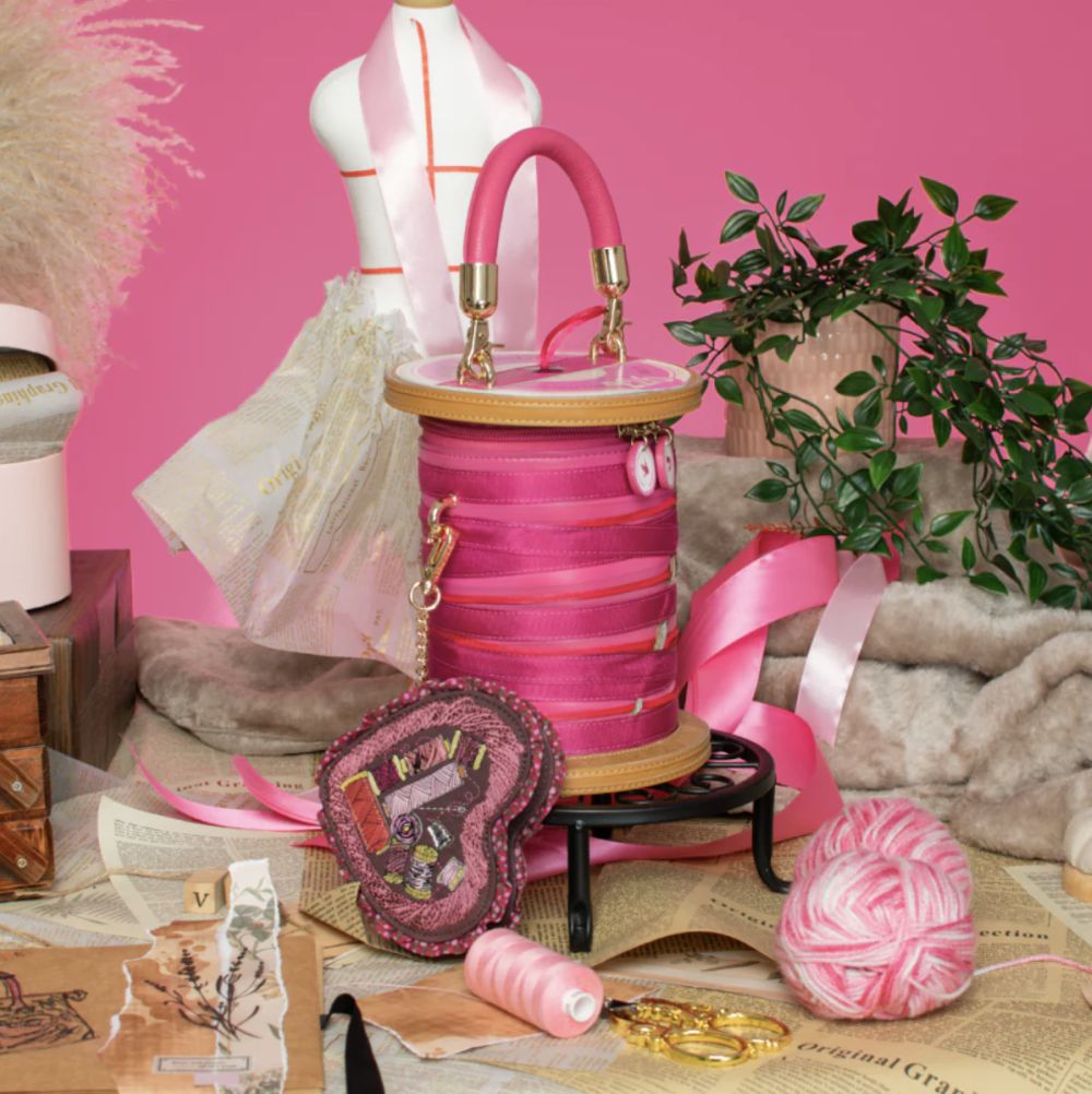 A pink spool-shaped handbag with a top handle, gold-tone hardware, and a quilted floral front, displayed on a small black stand in a styled craft room scene with pink yarn, thread, ribbon, and a mannequin torso against a pink backdrop.