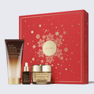 Estée Lauder holiday gift set featuring a red gift box with gold snowflake details, alongside a cream-colored skincare bottle, a small amber dropper bottle, and a gold-lidded cream jar, all arranged on a light gray background.