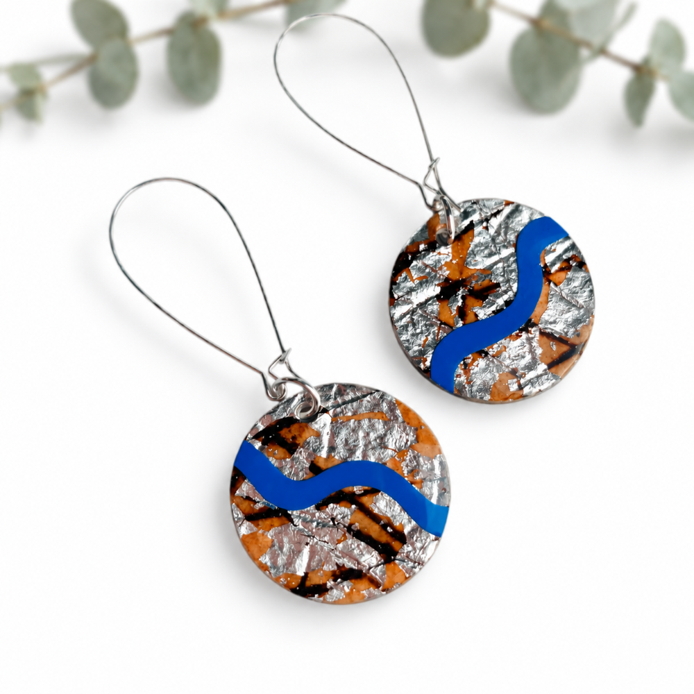 A pair of handmade round drop earrings featuring a textured silver foil background with a wavy blue stripe and orange/black abstract patterns, suspended from thin silver wire hooks on a white background with eucalyptus leaves.
