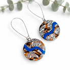 A pair of handmade round drop earrings featuring a textured silver foil background with a wavy blue stripe and orange/black abstract patterns, suspended from thin silver wire hooks on a white background with eucalyptus leaves.