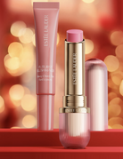 Estée Lauder Futurenist Blush-Maker heavy Cheeks tint and a matching lipstick in a ribbed pink and gold tube, displayed on a red surface with a warm bokeh background.