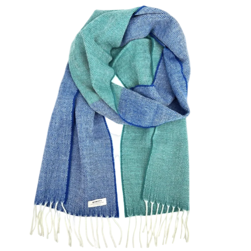 A woven scarf with a herringbone pattern in shades of blue and teal, finished with cream fringe and a small white label near one end, shown on a plain white background.