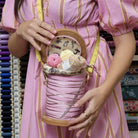 Person holding a pink spool bag with yarn and knitting needles, surrounded by fabric rolls.