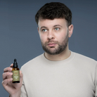 A man with short dark hair and a beard wearing a beige knit sweater, holding up a small amber glass dropper bottle labeled 'Shaving oil' against a solid slate blue-gray background.