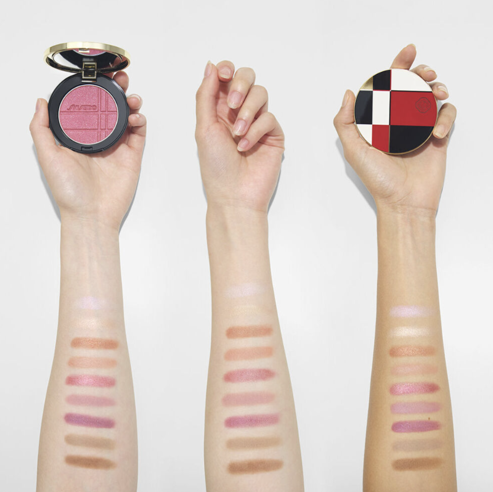 Three hands holding round makeup compacts against a white background, with swatches of the pressed powder applied on the forearms below each compact.