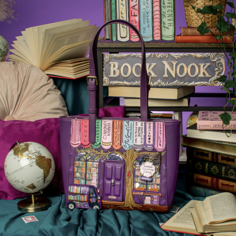 A purple handbag designed like a book nook with illustrated book spines and shelves, styled on a teal fabric surface with books, a globe.