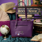 A purple leather tote bag with a front zip pocket and decorative text, styled on a teal fabric surface with books, a globe, and a purple background.