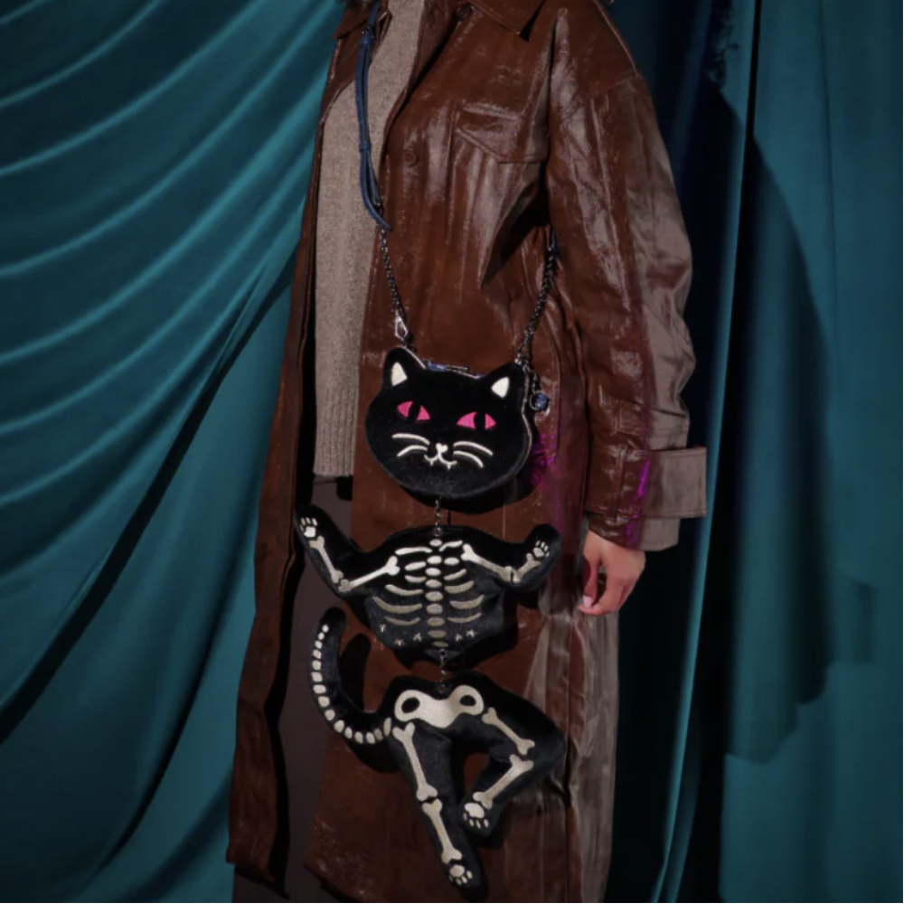 A model wearing a brown leather trench coat and beige sweater, carrying a black cat-shaped crossbody bag with a skeleton body design, set against a dark teal draped curtain background.