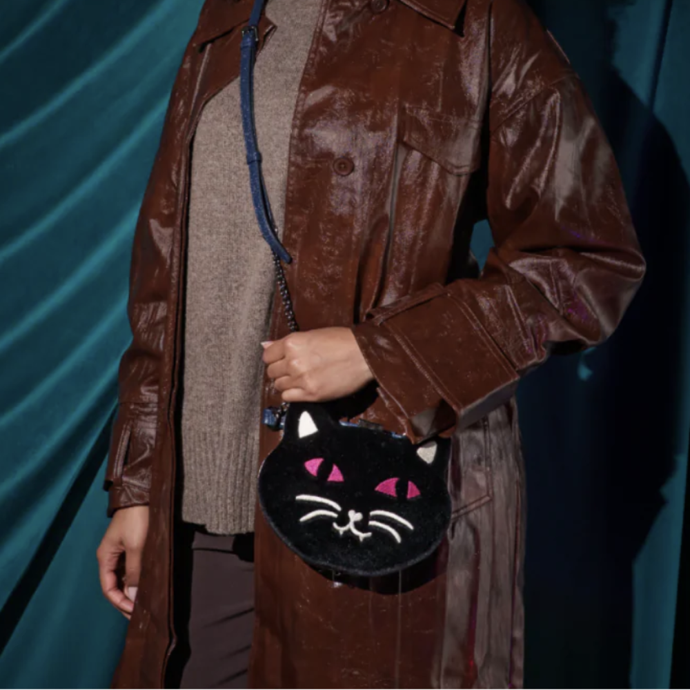 A person wearing a brown faux-leather trench coat over a beige knit sweater, holding a small black cat-face crossbody bag with pink embroidered eyes and white facial details, set against a dark teal draped backdrop.