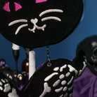 A close-up of a black velvet cat-shaped hanging bag with embroidered white facial features and pink eyes, shown against a blue background with other dark decorative items partially visible.