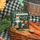 A small quilted dark green wallet with a whimsical embroidered design of a fox in a dress and the text 'The Farmhouse Pantry', styled in a rustic basket with carrots on a green-and-white checkered blanket.