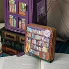 A purple zip-around wallet with a bookshelf illustration and a small brown coin purse placed in front of it, styled with old books and an open book on a soft surface.