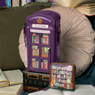 A purple phone-shaped clutch bag with a bookshelf window design and a small matching zip pouch placed in front, styled on books with an open book and cushion in the background.