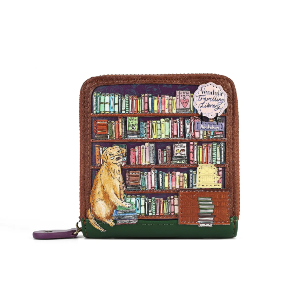A zip-around wallet with a brown leather border and a colorful printed illustration of a dog sitting in front of a bookshelf. The wallet has a small purple zipper pull and a decorative paper tag that reads 'Vendula Travelling Library'.