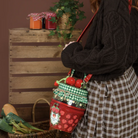 A woman wearing a black cable-knit sweater and brown-and-white plaid skirt carries a small red strawberry-themed shoulder bag with green gingham accents, standing in front of a rustic wooden crate against a brown background.