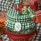 A close-up of a decorative strawberry jam jar bag with a red zipper around the base, covered by a green-and-white gingham fabric lid. A woven twine band holds a round white tag reading 'The Farmhouse Pantry'.