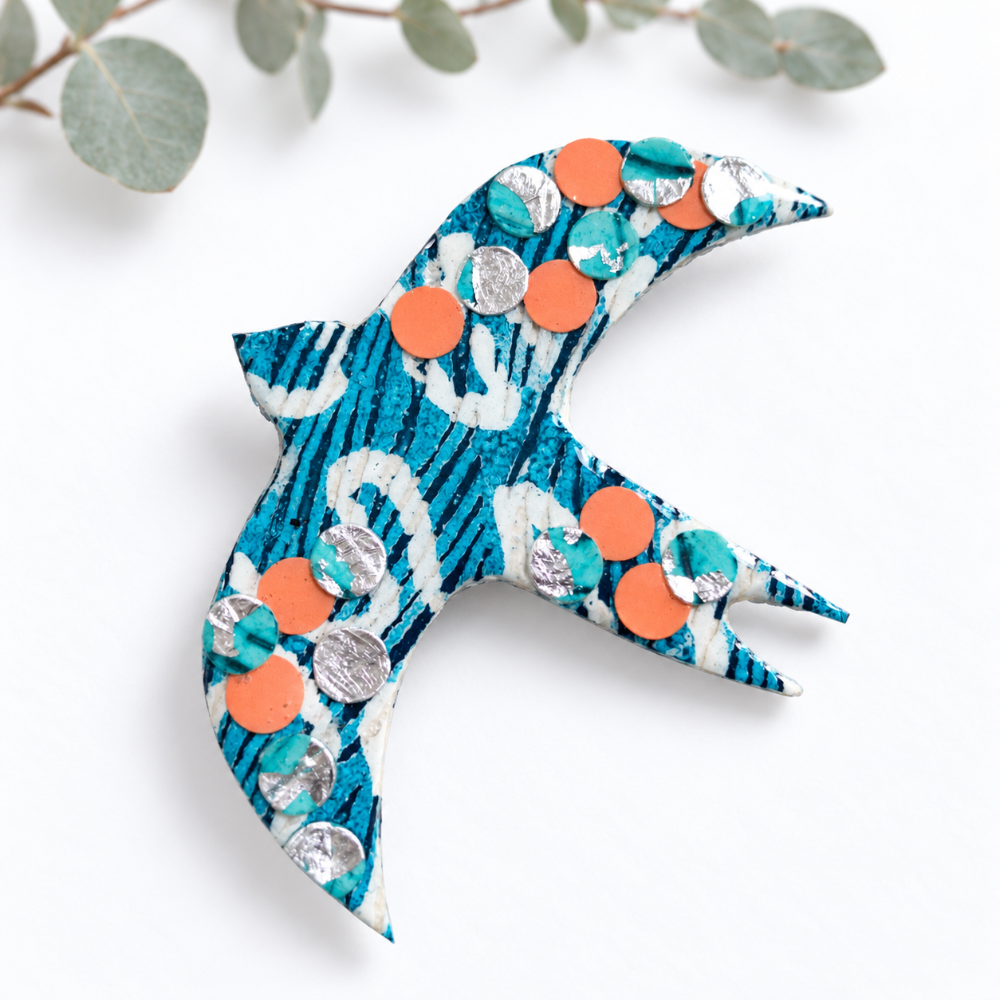 A bird-shaped decorative brooch or pin made from patterned fabric, decorated with orange and silver circular embellishments, photographed on a white background with eucalyptus leaves at the top.