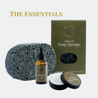 A grooming set labeled 'THE ESSENTIALS' featuring a dark charcoal porous shaving sponge, an amber dropper bottle of shaving oil, a tin of honey and tallow balm with its lid off, and an olive green box of BrownBull Infused Soap Sponge, arranged on a light gray background.