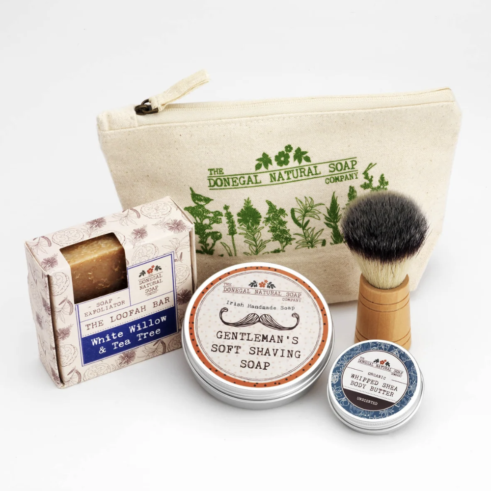A shaving gift set from The Donegal Natural Company on a white background, including a canvas zip pouch, boxed soap, two shaving soaps in tins, and a shaving brush.