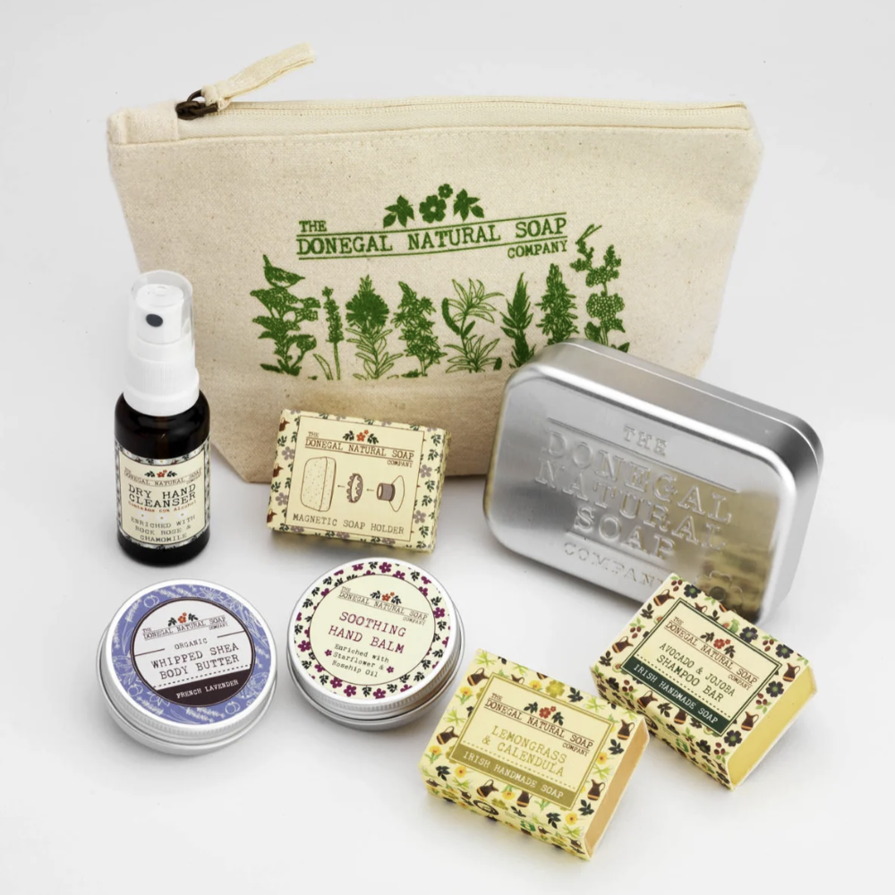 A set of natural soap products from The Donegal Natural Soap Company arranged on a white background, including a canvas zip pouch, a small dark bottle, a silver tin, and several wrapped bars and tins in different floral and botanical patterns.