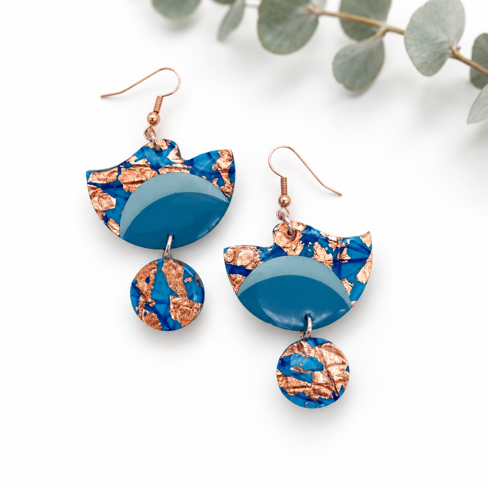 A pair of drop earrings with copper-toned fishhook wires, featuring a large semi-circular top piece and a dangling round charm. The main body of the earrings has a blue and copper foil crackle pattern with a solid light blue semi-circular center.